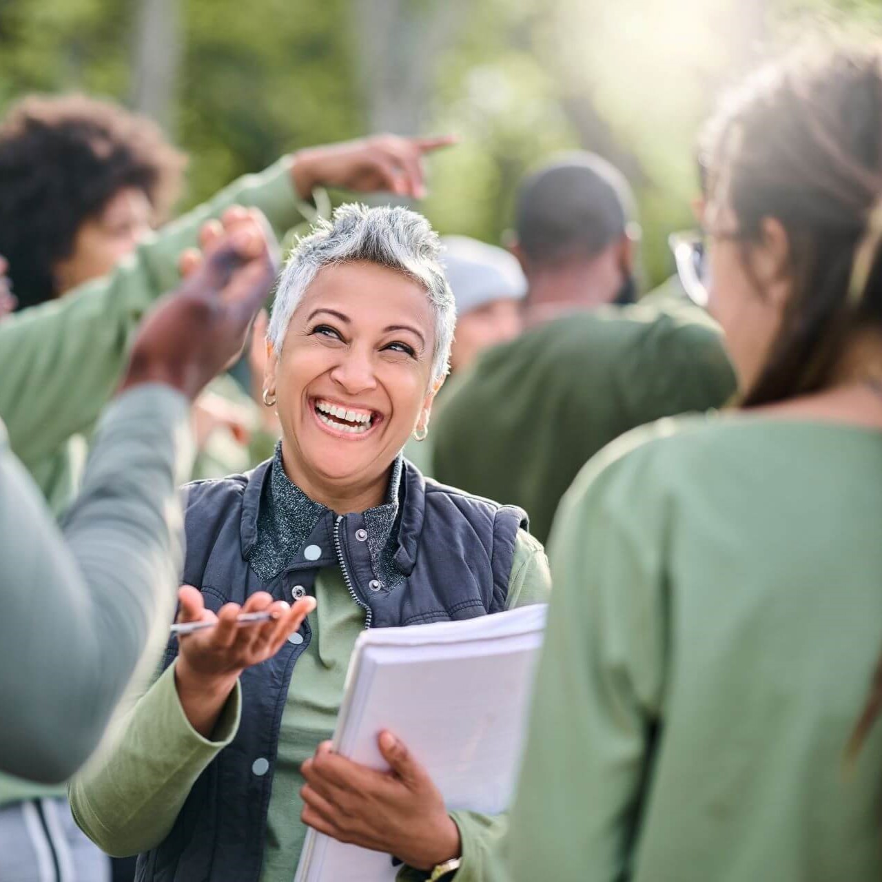Woman holding notepad, engaging happily with others in outdoor setting.